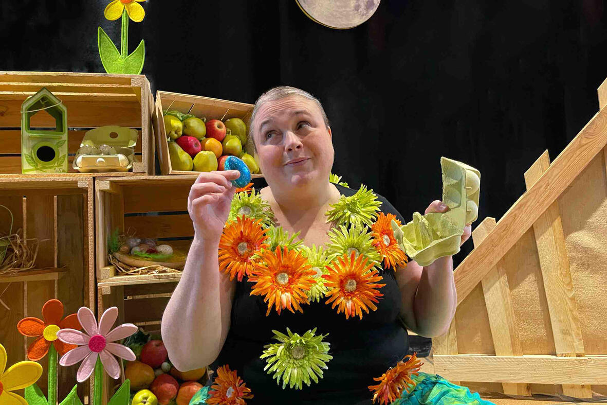 Spectacle j'me cache ! Scène de spectacle : Aurore conteuse interagit avec ses personnages et accessoires dans un décor en bois et fleurs.